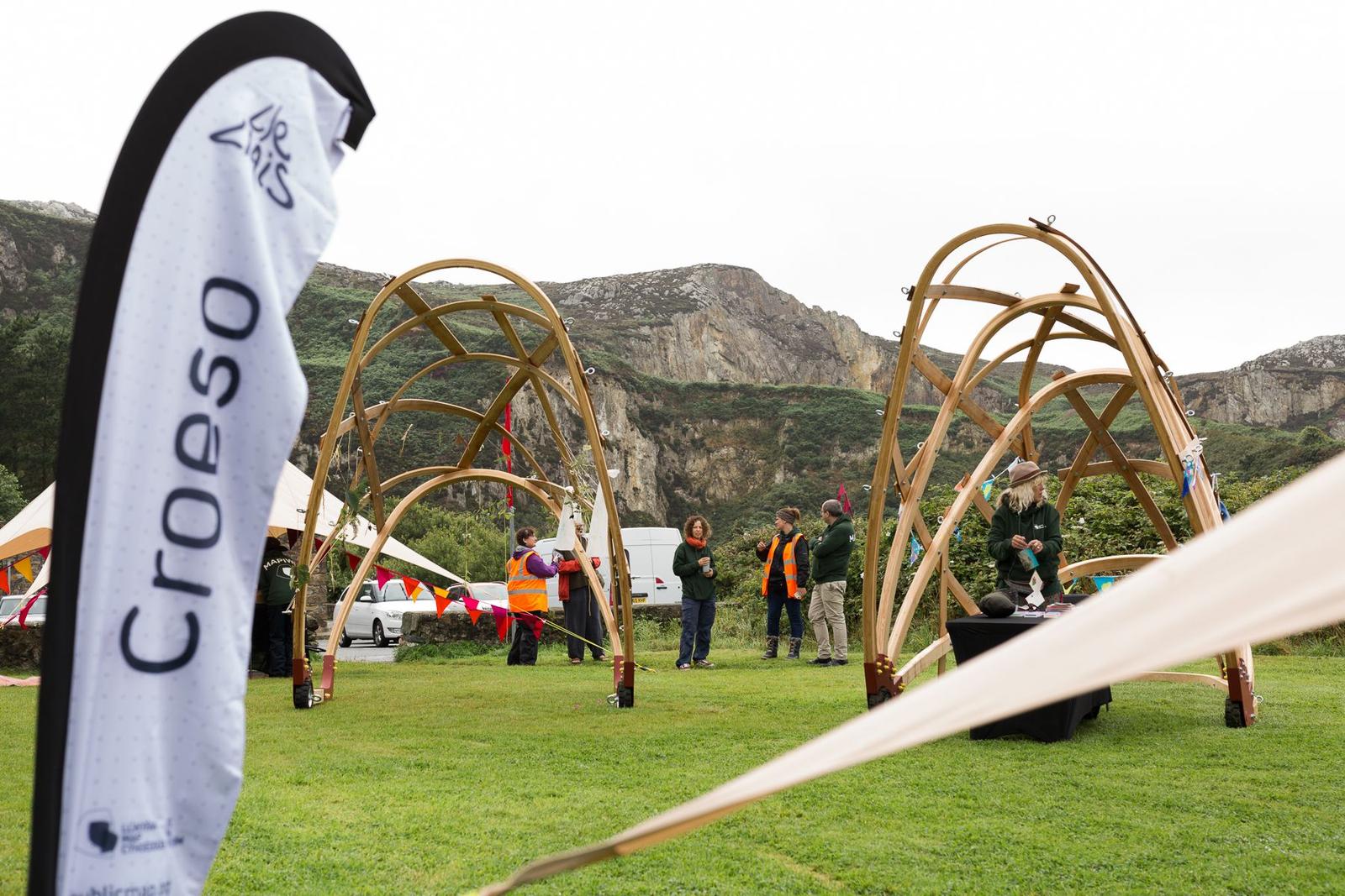 A view of a popup event space. Set in nature, tensile canopies and bentwood mesh structures sit with a mountain behind. In the foreground a branded flag with the Welsh word for 'welcome' - 'Croeso' is printed.